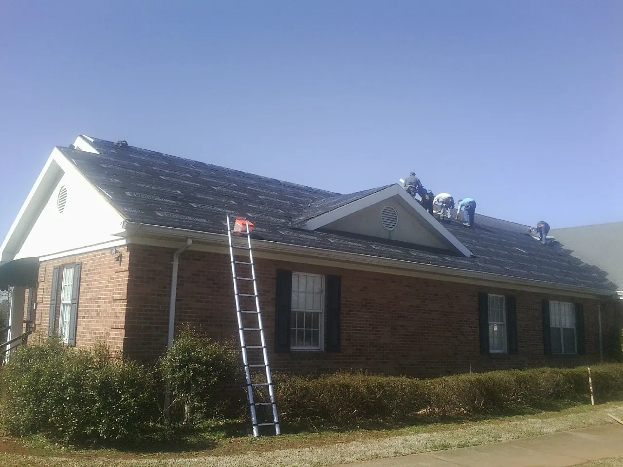 Crew tearing shingles off a brick ranch roof with a ladder set — Metal Roof Repair work by our crew in Hazlehurst
